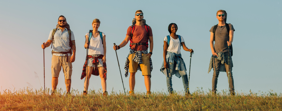 Five hikers wearing backpacks and carrying hiking poles standing side by side in the sunset smiling.